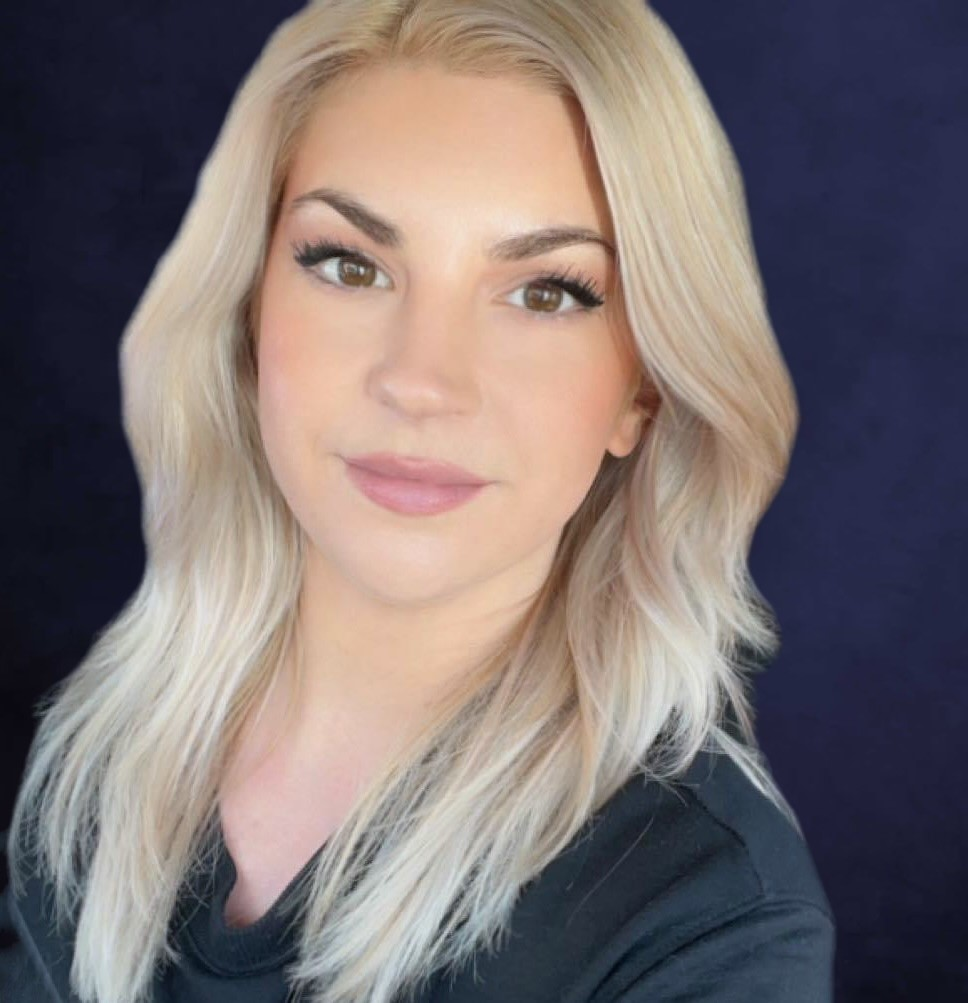 A smiling portrait of Eva Garcia Sizemore, the owner of Ultimate Reading Glasses. A young woman with blonde hair and auburn eyes, wearing a forest green shirt.