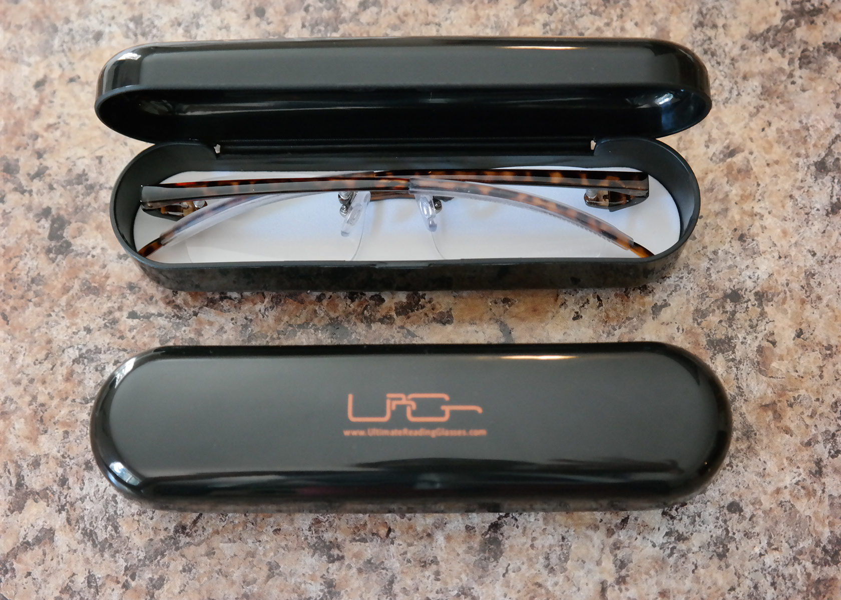 The Black Ultimate Reading Glasses hard case with tortoise reading glasses inside, resting on a table.